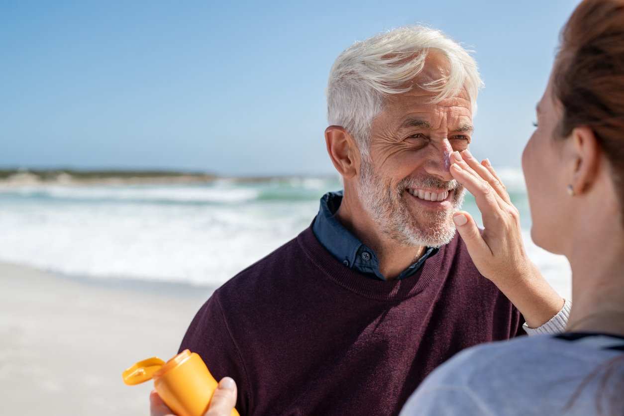Frau trägt einem älteren Mann am Strand Sonnencreme auf die Nase auf