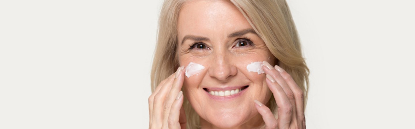 Hyaluronsäure: Lächelnde Frau mit Fältchen und Linien im Gesicht cremt ihre Wangen mit Sonnencreme ein 