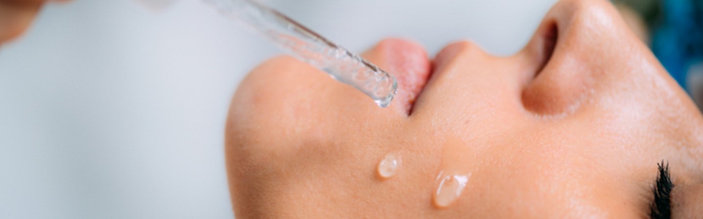  Einer Frau mit geschlossenen Augen werden zwei Hyaluronsäure-Serum-Tropfen auf die Wange geträufelt