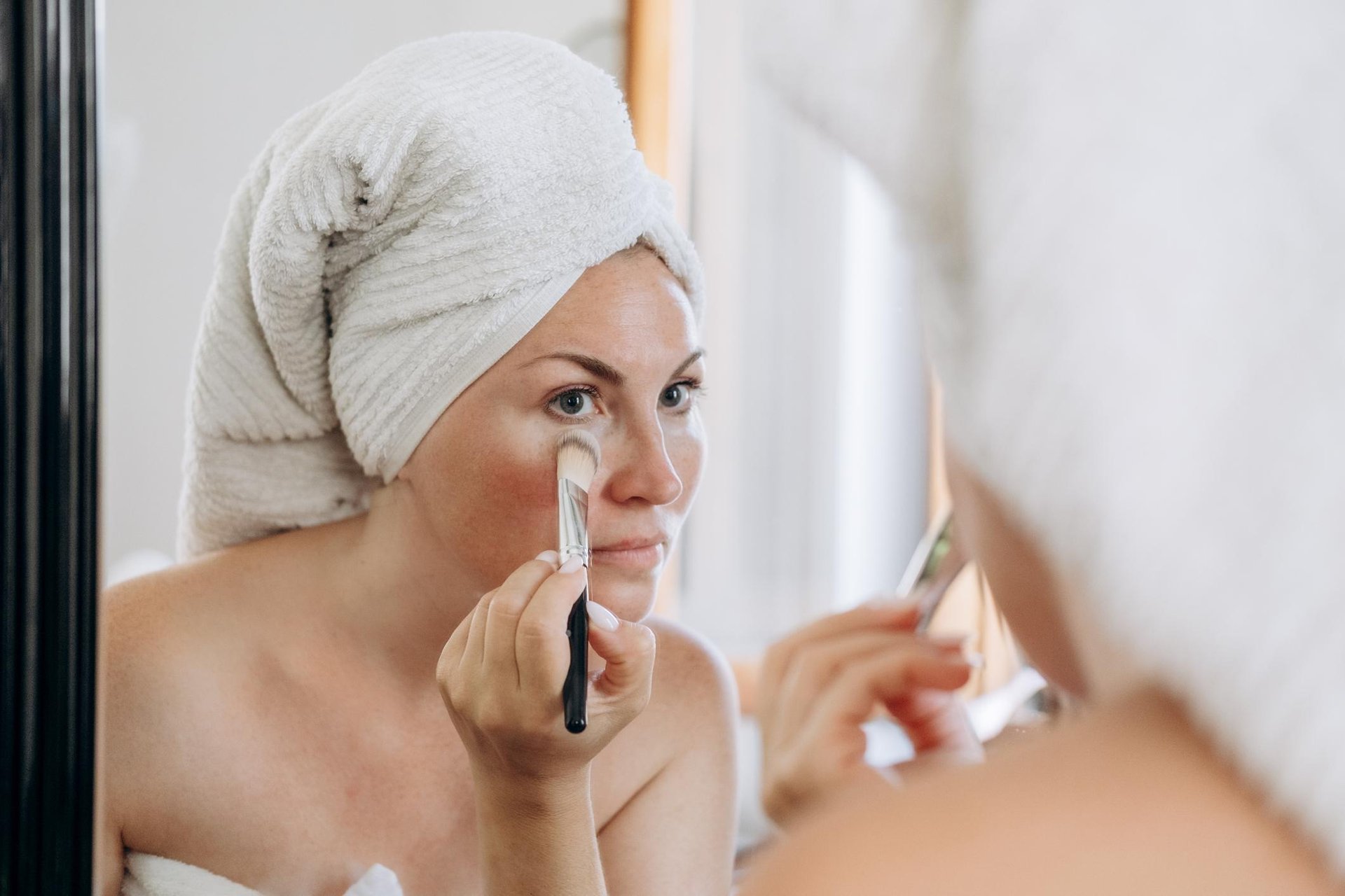 Frau mit einem Handtuch auf dem Kopf trägt mit einem weichen Pinsel Make-Up unter den Augen auf
