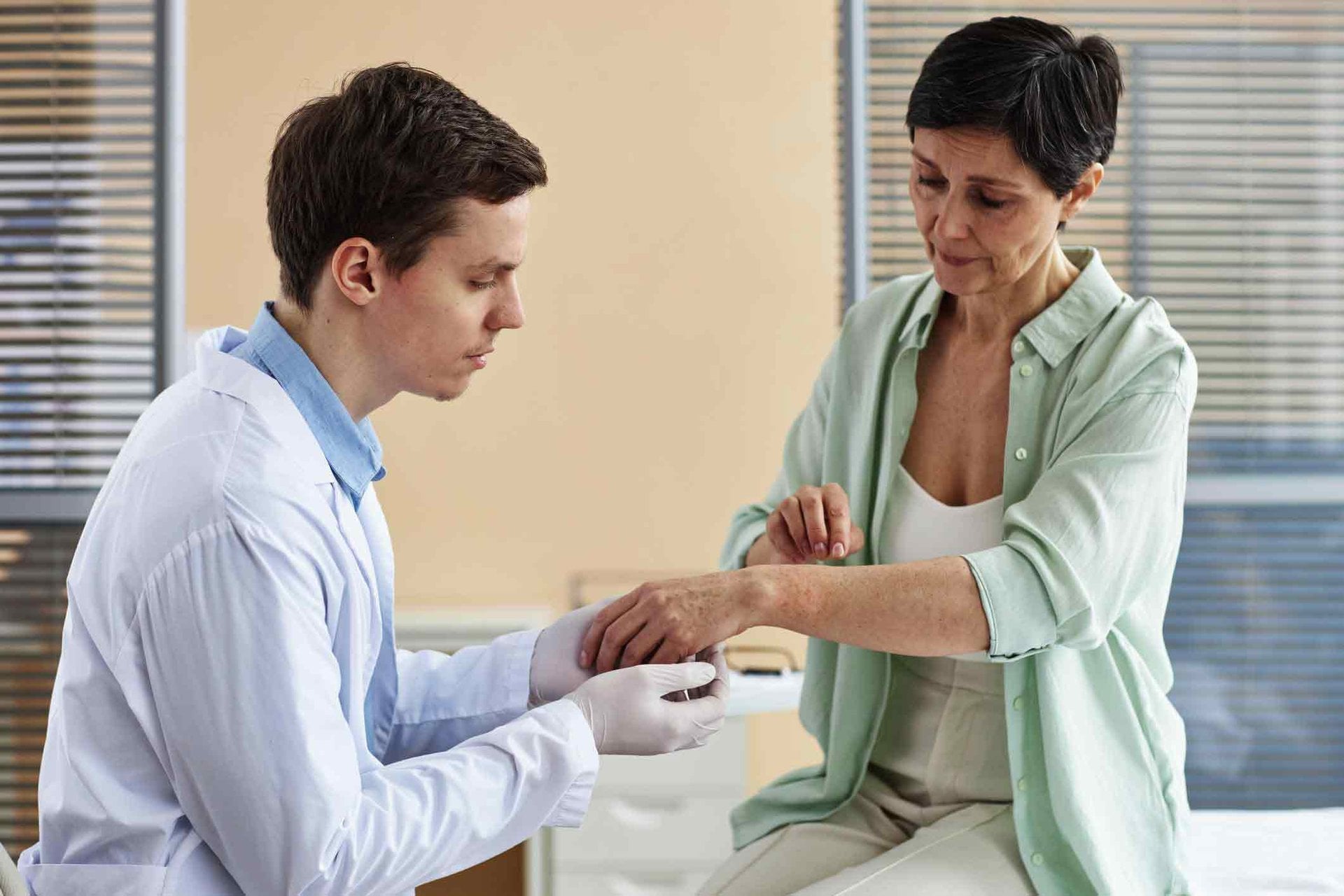 Arzt hält den Arm einer ihm gegenüber sitzenden Patientin, diese zeigt mit ihrer anderen Hand auf ihre Rötungen am Handgelenk