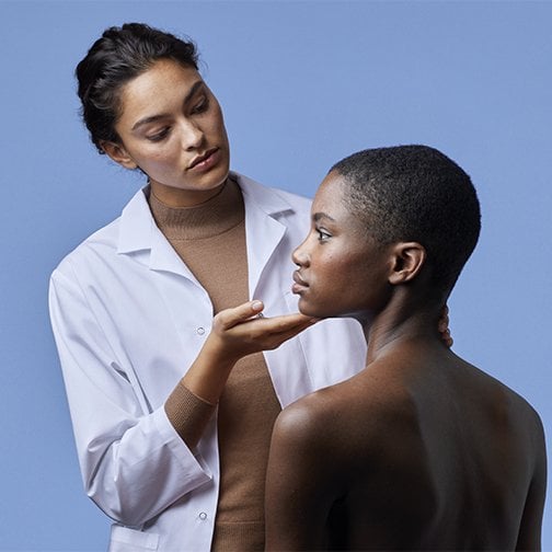 Une dermatologue tient le visage d'une patiente et le tourne sur le côté pour examiner sa peau.