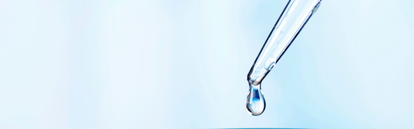 Eine klare Pipette mit einer herabhängenden Wassertropfen-Spitze vor einem unscharfen, hellblauen Hintergrund. Symbolisiert Präzision und Reinheit.