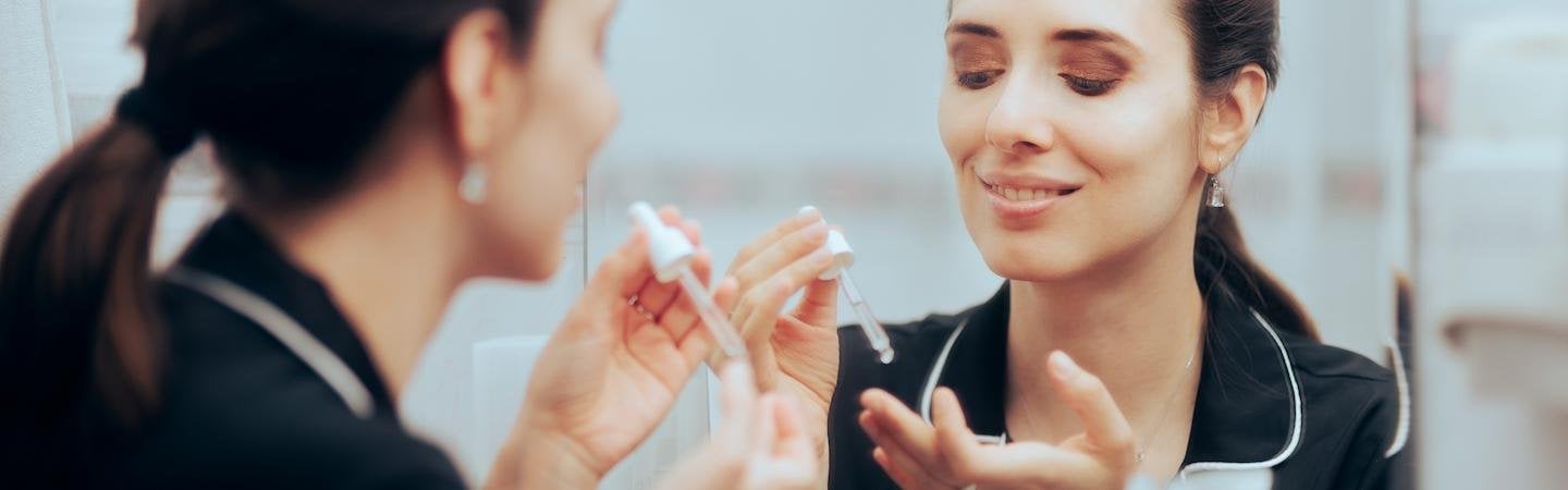 Frau steht vor einem Spiegel und trägt sich mit einer Pipette ein Serum auf die Finger auf.