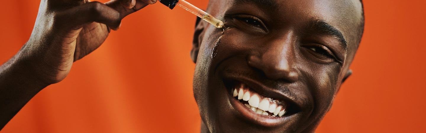Mann vor orangem Hintergrund lächelt breit in die Kamera, während er sich mit einer Pipette ein Serum ins Gesicht aufträgt.