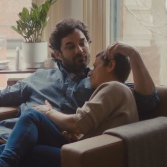 Un couple est assis, détendu, sur un canapé marron dans une pièce confortablement éclairée, avec une plante d'intérieur en arrière-plan, près d'une fenêtre.
