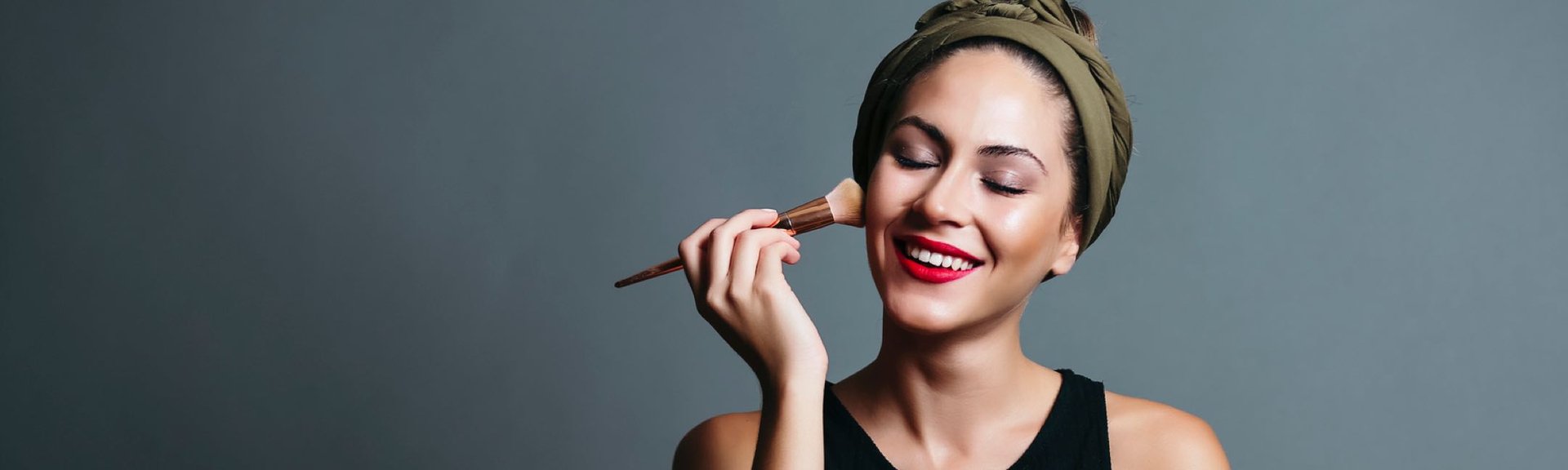 Lächelnde Frau mit olivgrünem Haarband, hält Schminkpinsel zum Auftragen von Make-up.