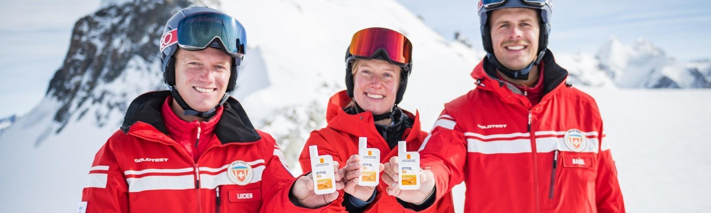 Zwei Männer und eine Frau in roter Skibekleidung und Helmen vor einer bergigen Schneelandschaft. Alle drei halten ein ANTHELIOS Produkt in der Hand und strecken es zur Kamera.