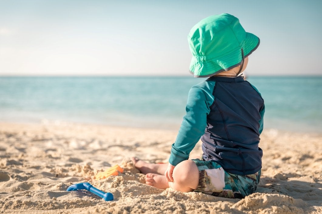 Kleinkind mit langer Kleidung und Hut in der Sonne am Strand